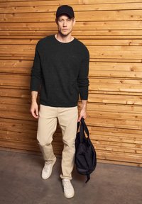 Homme portant une casquette noire, un pull sombre, un pantalon beige et des baskets blanches marchant à l'intérieur, tenant un sac à dos noir contre un mur en bois.