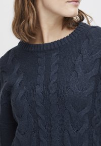 Mörkblå stickad tröja med kabelmönster, rundad halsringning och textilstruktur. Synliga stygn och ribbade muddar.