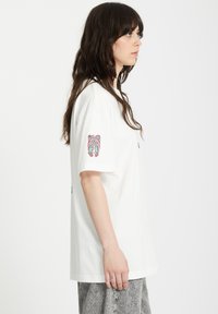 Camiseta blanca de algodón con mangas cortas, que presenta un estampado gráfico colorido en la manga izquierda y un corte relajado. Parte inferior gris con estampado.