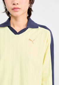 Polo shirt Puma en jaune clair avec texture côtelée, col bleu marine et logo brodé sur la poitrine. Présente un design en col en V.