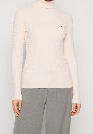 Kobieta w jasnoróżowym, teksturowanym swetrze z golfem i szarych szerokich spodniach na jednolitym tle.