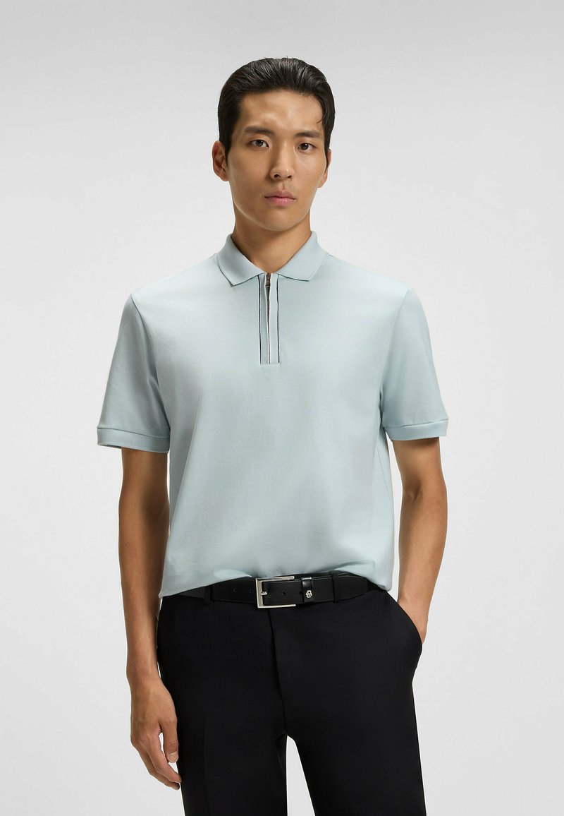 Ljust blå polo med dragkedja i kragen, korta ärmar och svarta sömnadsdetaljer, parat med svarta byxor och ett bälte.