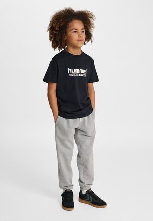 Barn med krøllet hår står og kigger fremad iført sort "hummel" skjorte, grå joggingbukser og sorte sneakers med grønne accenter.