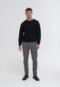 Must piloot kõrge kraega mustad sviitrid, hallid kohandatud püksid ja mustad nahkkingad. Mudel seisab ühetoonilise valge tausta ees.