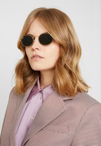 Occhiali da sole neri rotondi con sottili montature in metallo dorato; giacca a quadri marroni e bianchi con texture sopra una camicia di un leggero color viola.