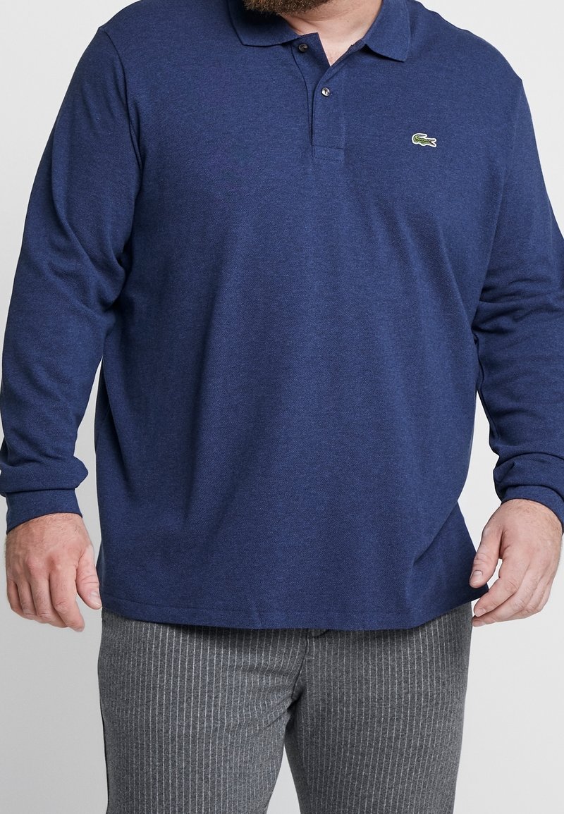 Marineblaues Langarm-Polohemd aus strukturiertem Stoff, mit einem Drei-Knopf-Verschluss und einem kleinen grünen Krokodil-Logo auf der Brust. Kombiniert mit gestreiften Hosen.