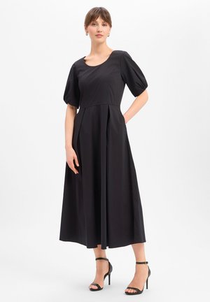 Kobieta w czarnej sukience midi z bufkami i plisami, połączona z czarnymi szpilkami z paskiem wokół kostki, stojąca na białym tle.