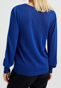 Blå stickad tröja med långa puffärmar, ribbad nederdel och en glänsande textur. Bakvyn visar en rundad halsringning och en avslappnad passform.