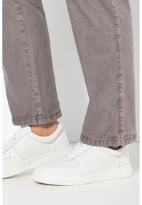Weiße Leder-Sneaker mit durchbrochenem Design, kombiniert mit grau verwaschenen Hosen. Die Sneaker verfügen über flache weiße Schnürsenkel und eine Gummisohle.