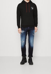 Černá mikina s oranžovými šňůrkami a logem na hrudi, doplněná o modré distressed džíny a černé tenisky, vytvářející neformální outfit.