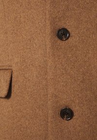 Bruine wollen jas met een zachte textuur, voorzien van twee zwarte knopen en een klepzak aan de linkerkant.