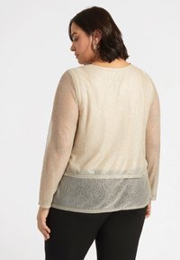 Maglietta a maniche lunghe in beige chiaro, caratterizzata da un motivo floreale testurizzato, fili lucidi e un pannello posteriore trasparente, abbinata a pantaloni neri.