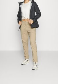 Svart lättviktsjacka med huva, beiga byxor och vita sneakers. Påfallande dragkedjedetaljer och en knuten vit tröja under jackan.