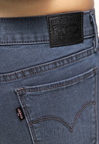 Vaqueros de mezclilla azul oscuro con una parche de cuero negro con un logo en relieve y bolsillos traseros cosidos, acentuados con hilo de contraste.