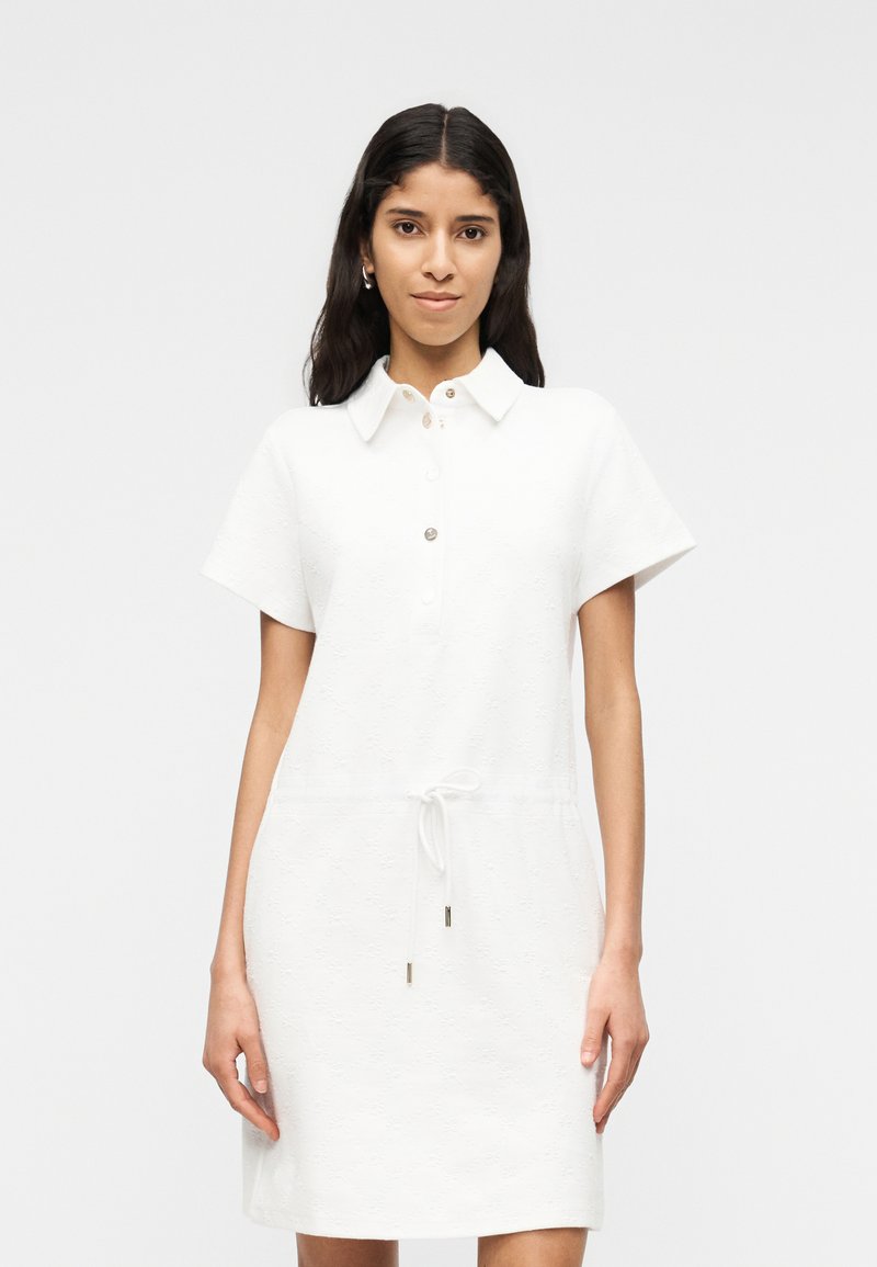 Femme aux longs cheveux noirs portant une robe blanche à manches courtes avec un col, des boutons et une ceinture nouée, debout devant un fond uni.