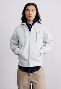 Sweatshirt gris à fermeture éclair en tissu doux avec une capuche à cordon et un logo jaune sur la poitrine. Porté par-dessus une chemise en flanelle bleue à carreaux.