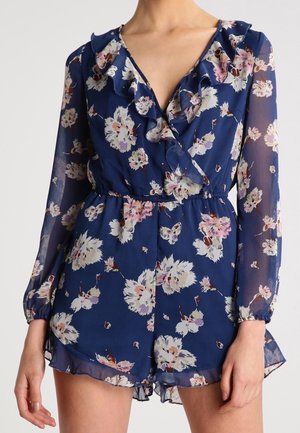 Kvinde iført en blå blomsterprintet langærmet romper med flæsekant ved halsen og elastisk talje, stående foran en ensfarvet baggrund.
