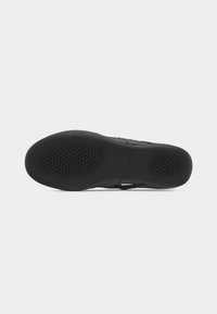 Semelle d'une chaussure noire avec des zones de grip texturées sur l'avant-pied et le talon, présentée à plat sur un fond uni clair.