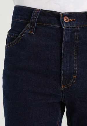 Close-up af mørkeblå denimjeans i taljen med synlig knap, lynlås, bæltestropper og orange syninger.