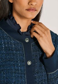 Nahaufnahme einer Person, die eine geknitterte marineblaue Bluse unter einer strukturierten marineblauen Jacke mit zwei metallischen Knöpfen anpasst.