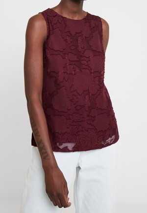 Bordeauxrotes ärmelloses Oberteil mit floraler, strukturierter Stickerei, mit rundem Ausschnitt und leicht geschwungenem Saum, kombiniert mit hellen Jeans aus Denim.