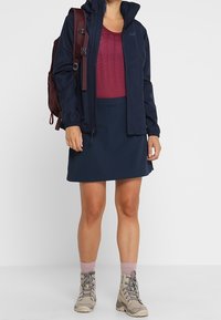 Marinfärgad vattentät jacka med huva, mörkröd andningsbar tröja och marinfärgad kjol. Beigefärgade vandringsskor med rosa strumpor. Brun ryggsäck med axelremmar.