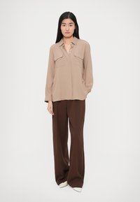 Cămașă bej cu mâneci lungi și două buzunare la piept, asortată cu pantaloni maro cu croială largă și pantofi cu toc alb, vârful ascuțit. Texturi netede pe întreaga îmbrăcăminte.