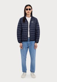 Navy puffer jacket with a zip front and quilted design. Worn over a white t-shirt, paired with blue jeans and white sneakers.
