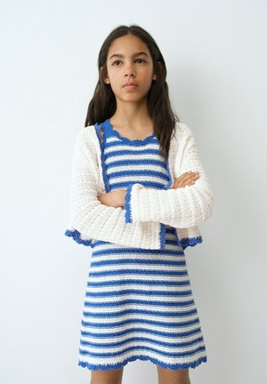 Fată cu păr lung și întunecat, purtând o rochie tricotată albastru cu dungi albe și un cardigan din tricot alb, stând cu brațele încrucișate.