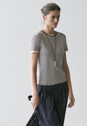 Ung kvinde iført en stribet kortærmet skjorte, sort nederdel og vedhængshalskæde, der holder en sort taske mod en ensartet lys baggrund.