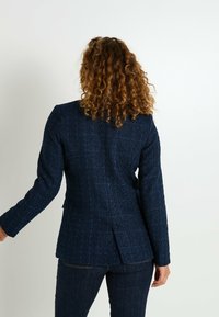 Blazer texturizado azul marino con un diseño ajustado, mangas largas y una abertura en la parte trasera. Hecho de un material tejido con una apariencia estructurada.