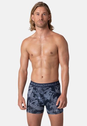 Shirtless man with shoulder-length blond hair wearing navy blue boxer briefs with a monkey and wave pattern, branded waistband visible.