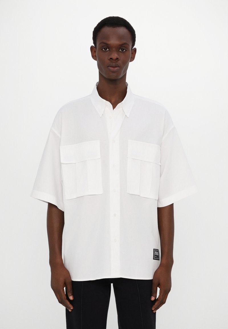 Witte kortek mouwenshirt met een button-down kraag, twee grote borstzakken en een patchlabel aan de onderzijde links. Soepele textuur.