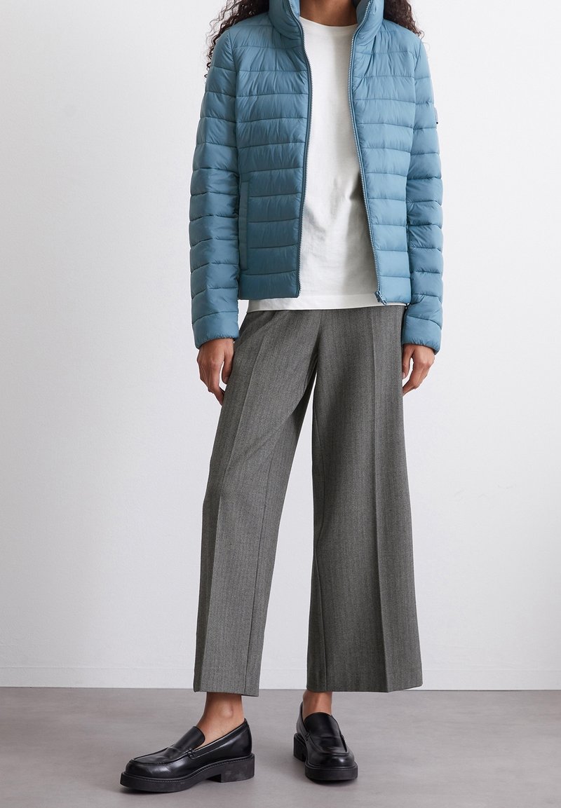 Helle blaue Steppjacke mit Stehkragen, strukturierten Einsätzen, weißem T-Shirt darunter, grauen weiten Hosen und schwarzen Slippern.