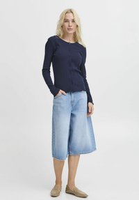 Námornícky pruhovaný cardigan s gombíkmi, skombinovaný s svetlomodrými voľnými džínsovými šortkami. Model má na sebe béžové šľapky.