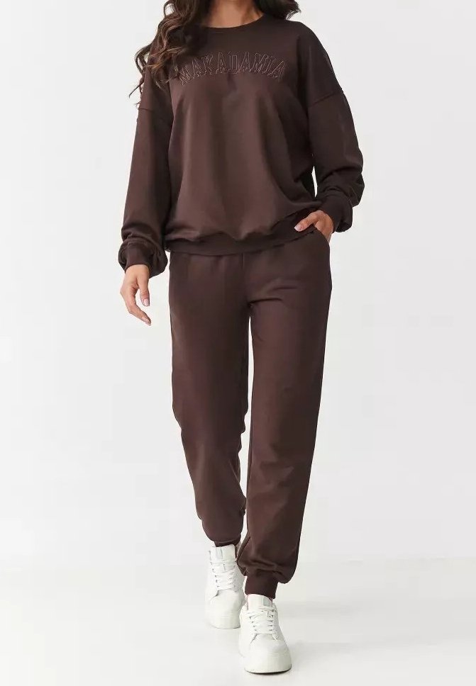 Femme portant un sweat-shirt marron foncé et un pantalon de jogging assorti avec des baskets blanches, marchant devant un fond uni.