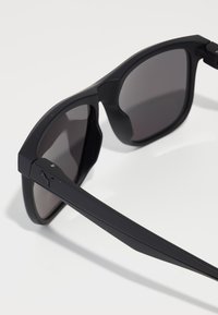 Ochelari de soare rectangulari negri cu rame mate și lentile închise. Caracteristici includ un logo distinctiv pe latură și un design elegant, ușor.