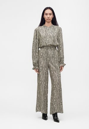 Femme debout portant un chemisier à manches longues imprimé léopard assorti et un pantalon large avec des bottines noires sur fond blanc.