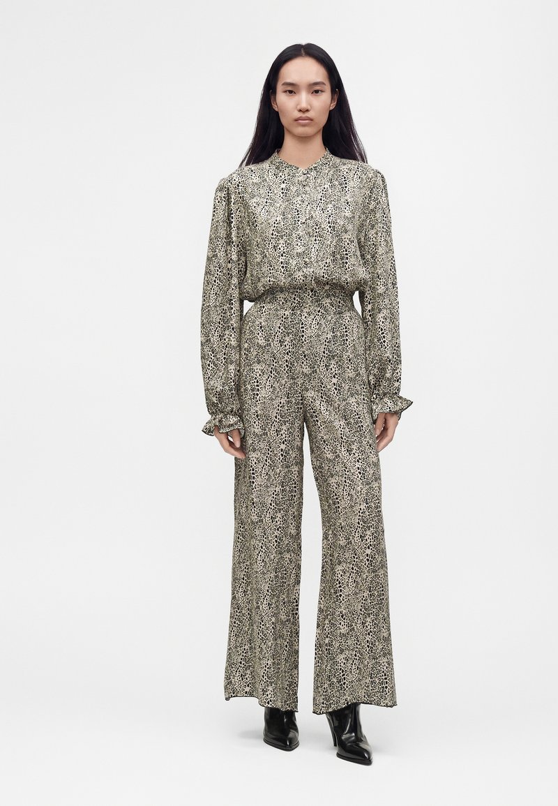 Femme debout portant un chemisier à manches longues imprimé léopard assorti et un pantalon large avec des bottines noires sur fond blanc.