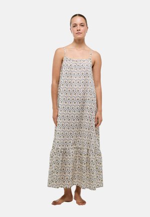Femme debout pieds nus vêtue d'une longue robe fleurie sans manches avec de fines bretelles sur un fond clair uni.
