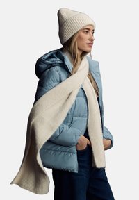 Chaqueta acolchada azul claro con capucha, llevada sobre una parte superior oscura; bufanda de punto acanalado en color crema colgada alrededor del cuello; textura suave.