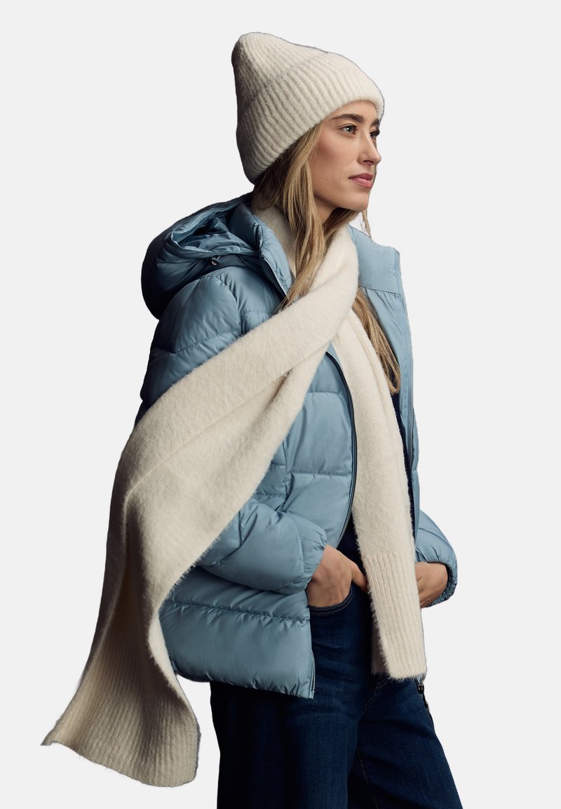 Chaqueta acolchada azul claro con capucha, llevada sobre una parte superior oscura; bufanda de punto acanalado en color crema colgada alrededor del cuello; textura suave.