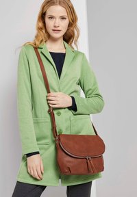 Borsa a tracolla in pelle marrone con chiusura a zip e dettaglio con nappina, abbinata a un blazer verde chiaro con texture.