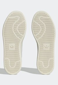 Skosulor i off-white med texturerad yta och hexagonalt mönster, med en central vågdesign och varumärkeslogotyp i hälområdet.