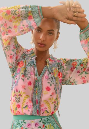 Blumenbluse aus transparenter Stoff mit pinkem Grundton und grünen sowie mehrfarbigen Akzenten. Rüschenkragen, Knopfverschluss und lange Ärmel.