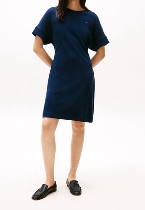 Vrouw staat met handen achter de rug, draagt een marineblauwe kortemouwige knielange jurk en zwarte loafers op een witte achtergrond.