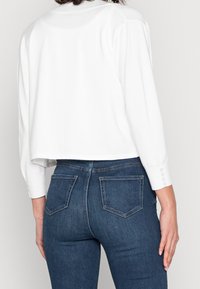 Biała krótka bluzka z długimi rękawami i guzikami, noszona z ciemnoniebieskimi dopasowanymi dżinsami z kieszeniami naszytymi.