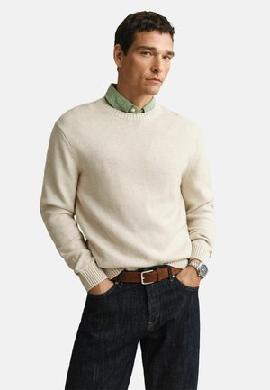 Mann trägt cremefarbenen Rundhalspullover über grünem Hemd mit Knöpfen, dunkle Jeans, brauner Gürtel und silberne Armbanduhr, steht mit den Händen in den Taschen.