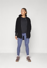 Black textured outerwear with a high collar, layered over a fitted black top, paired with light blue leggings and brown sneakers.