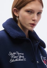 Tamsiai mėlynas paltas su pūkuotu apykakliu, puoštas baltais siuvinėjimais ir sagų užsegimu, eksponuojamas prieš šviesią fono spalvą.